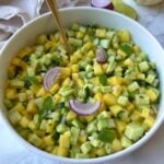 A bowl of diced mango, cucumber, and avocado salad garnished with red onion slices and mint leaves, with a gold spoon in the bowl.