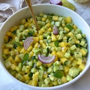 A bowl of diced mango, cucumber, and avocado salad garnished with red onion slices and mint leaves, with a gold spoon in the bowl.