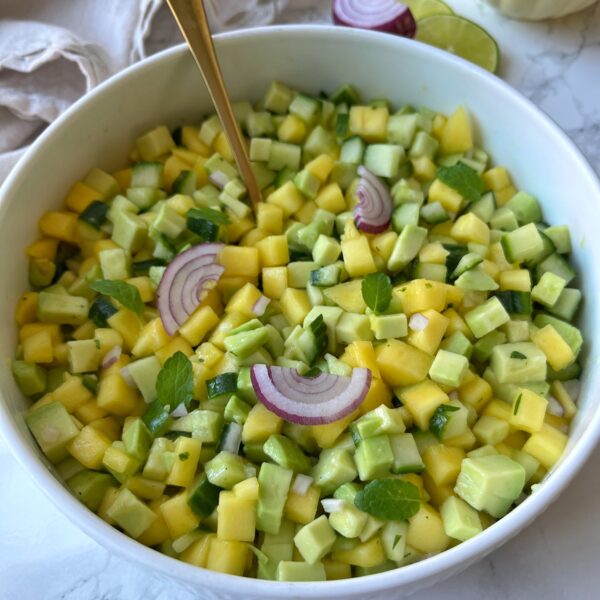 A bowl of diced mango, cucumber, and avocado salad garnished with red onion slices and mint leaves, with a gold spoon in the bowl.