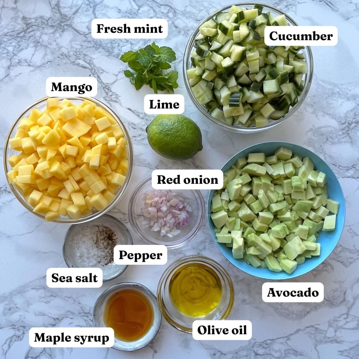 Bowls of diced mango, cucumber, avocado, red onion, fresh mint, a whole lime, plus olive oil, maple syrup, sea salt, and pepper are arranged on a marble surface.