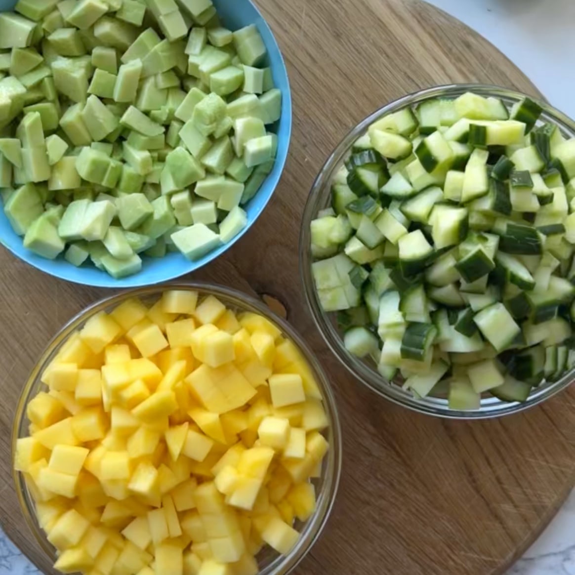 Three bowls with diced mango, avocado, and cucumber are placed on a wooden board.