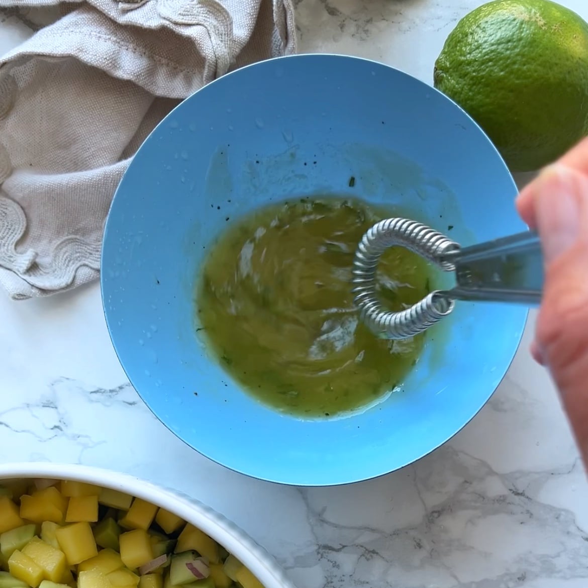 A hand whisking a green dressing in a blue bowl, with a lime, diced mango, and a cloth nearby on a marble surface.