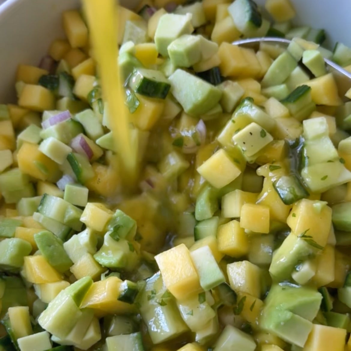 Close-up of an Avocado Mango Salad: diced avocado, mango, cucumber, and red onion in a bowl as a liquid dressing is poured over the fresh mixture.