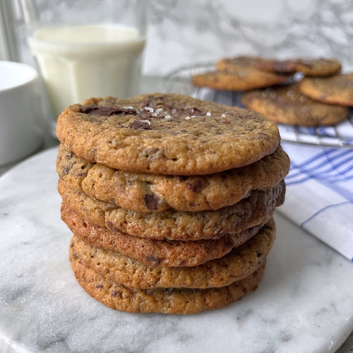 A stack of banana bread cookies sits on a marble surface, with a glass of milk and more cookies in the background.
