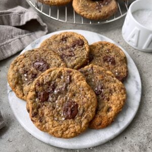 A plate of banana bread cookies topped with sea salt with a cooling rack of cookies in the background.