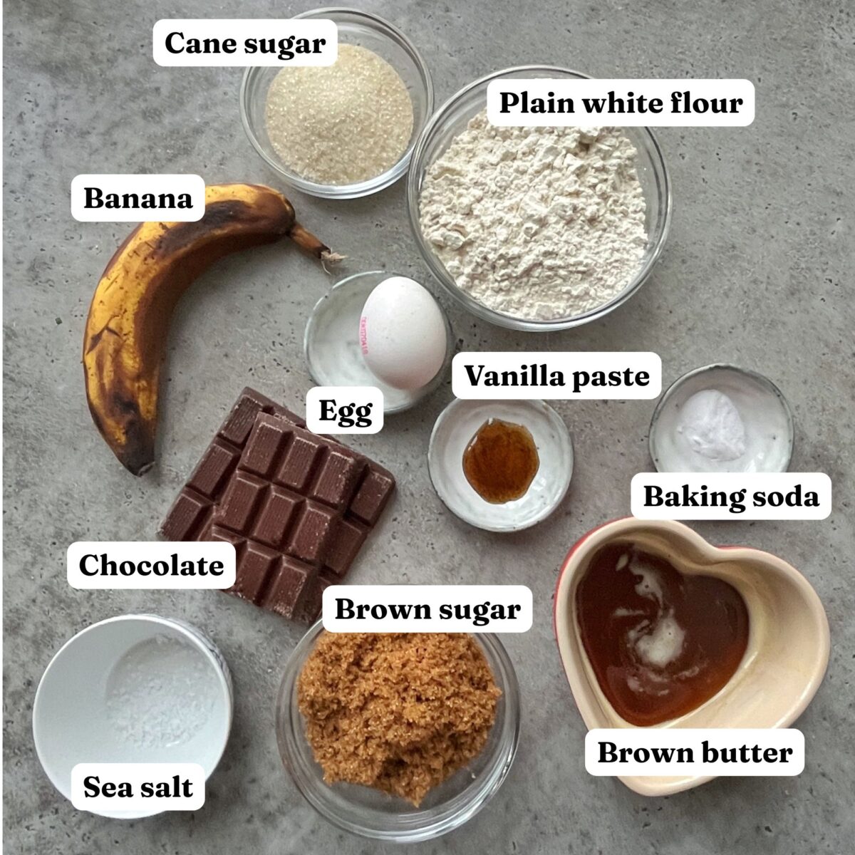 Banana bread cookie ingredients on a gray surface, including a banana, chocolate, egg, vanilla paste, brown and cane sugar, sea salt, plain flour, baking soda, and brown butter.