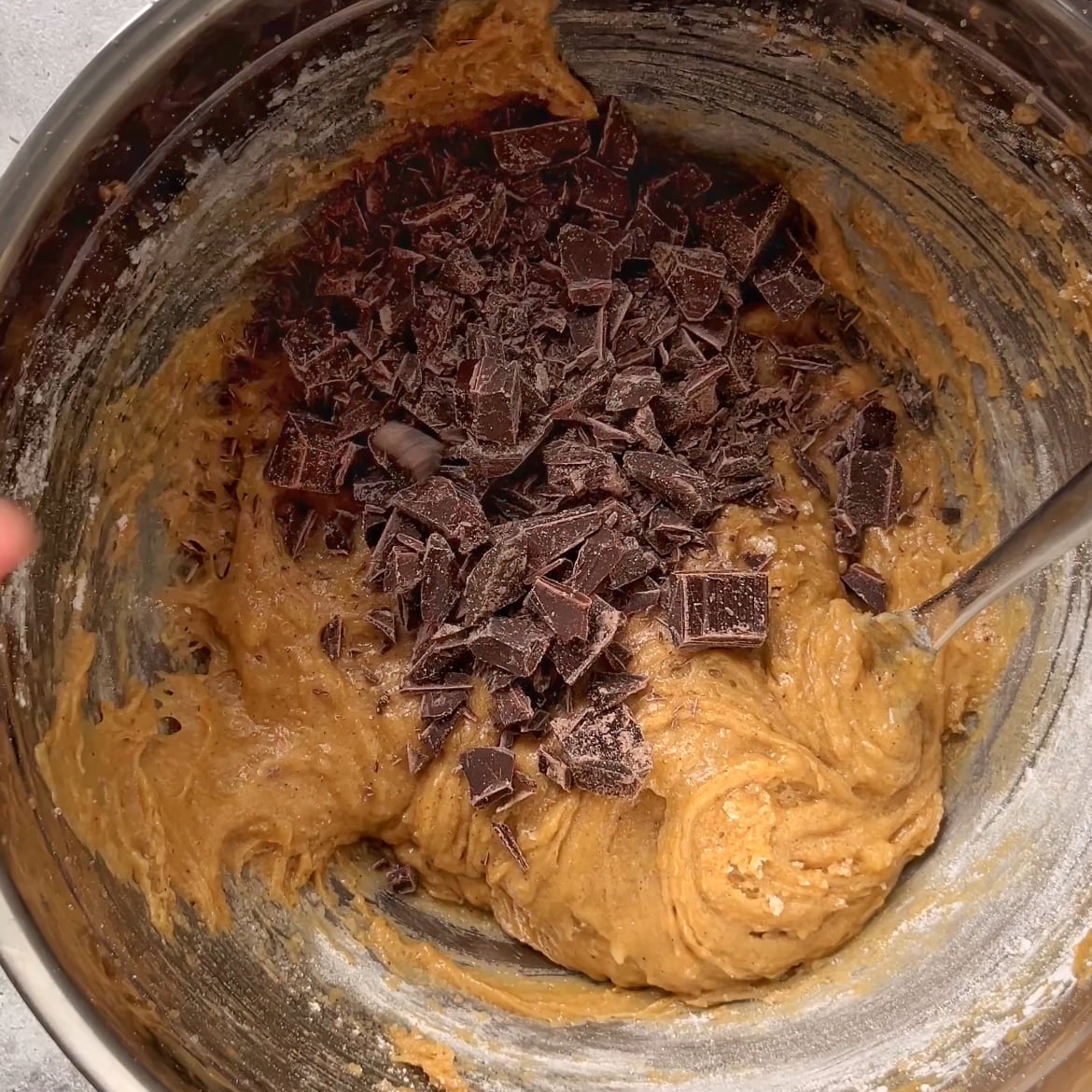 A metal bowl filled with cookie dough topped with chunks of chopped dark chocolate, being mixed with a metal spoon.