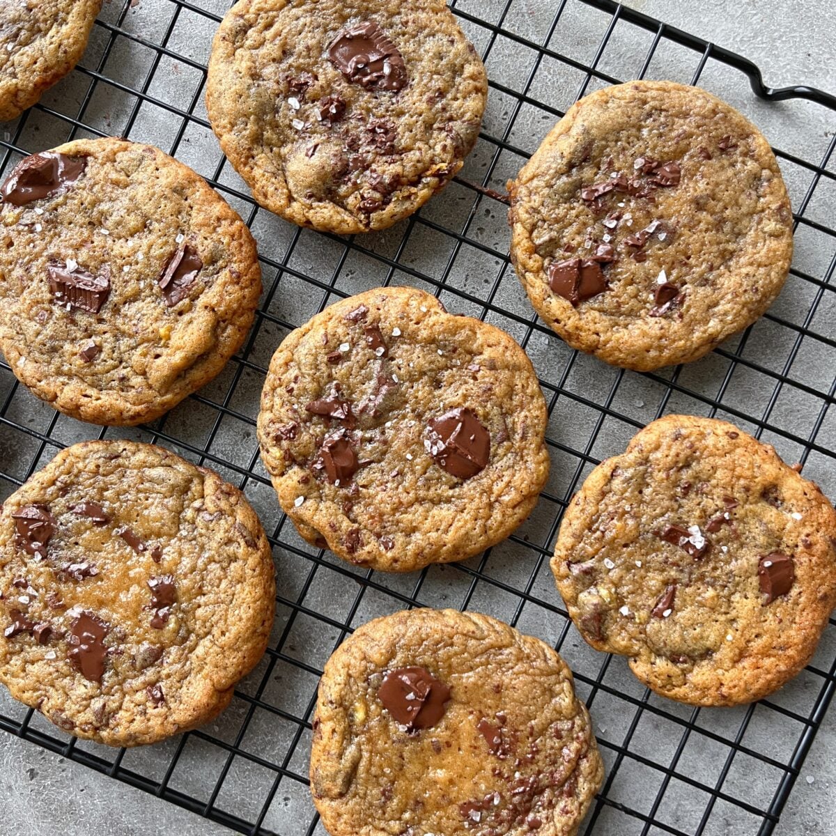 Eight chocolate chip cookies with visible chocolate chunks and sprinkled sea salt cooling on a black wire rack.