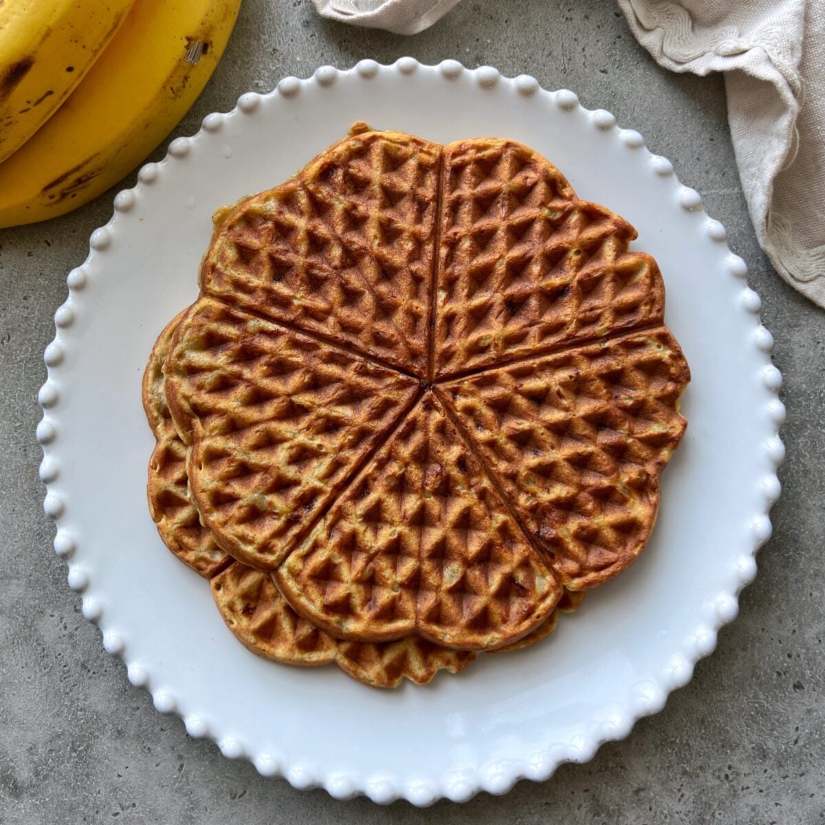 Two round waffles with six segments are served on a white plate with a beaded edge, set on a gray surface. Bananas and a napkin are visible nearby.