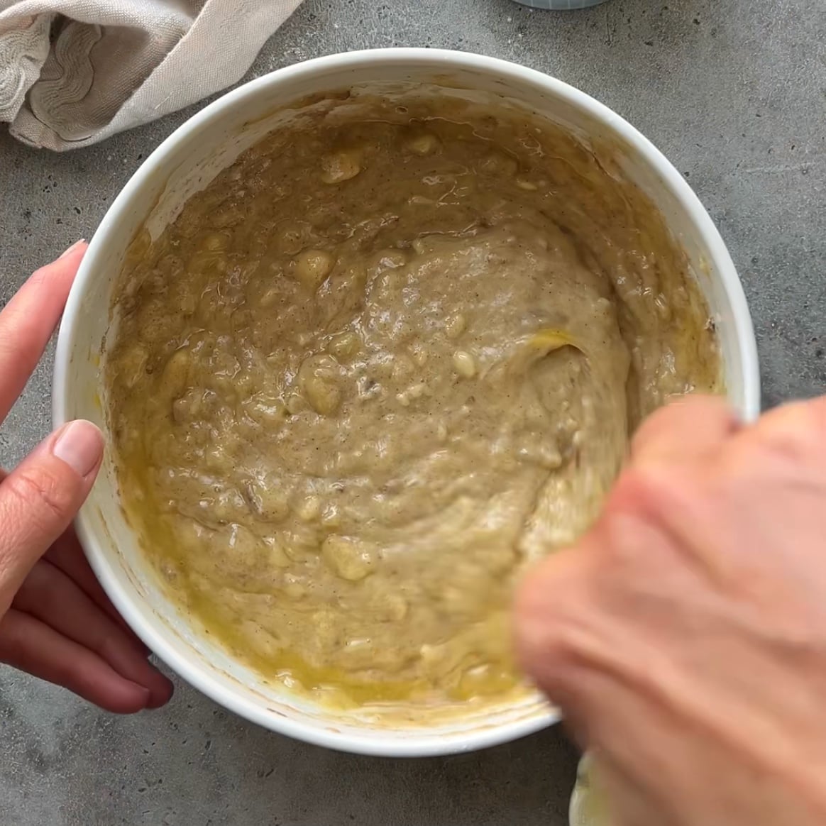 A hand mixes a banana waffle batter in a bowl with a fork. A cream colored tea towel is visible in the background.