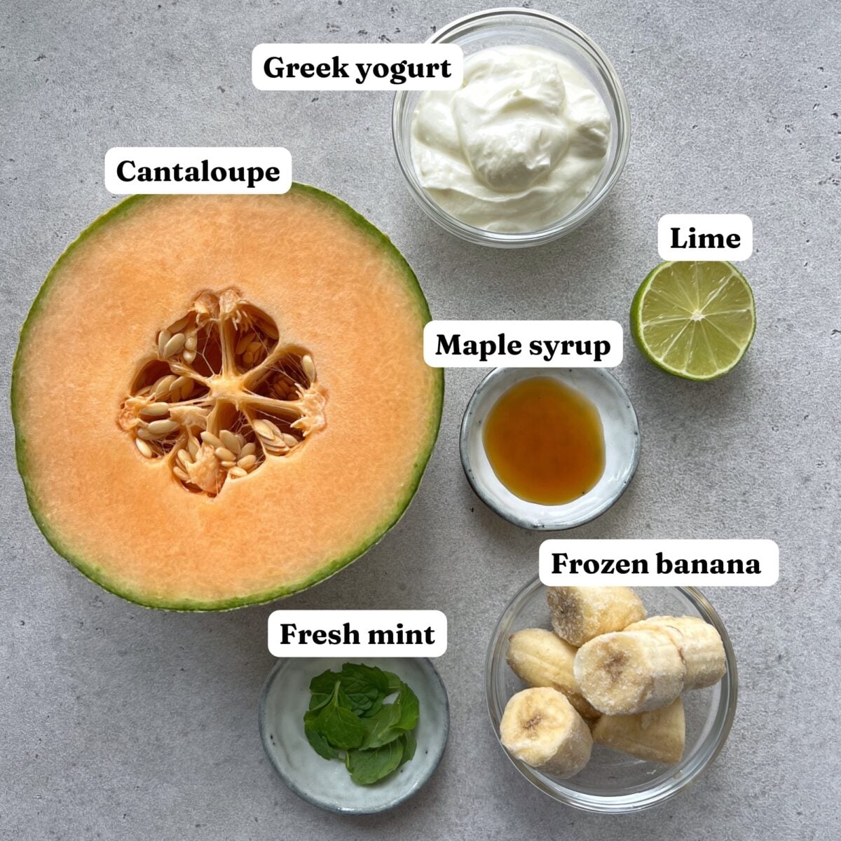 A halved cantaloupe, Greek yogurt, frozen banana, maple syrup, fresh mint leaves, and a halved lime are arranged on a gray surface.