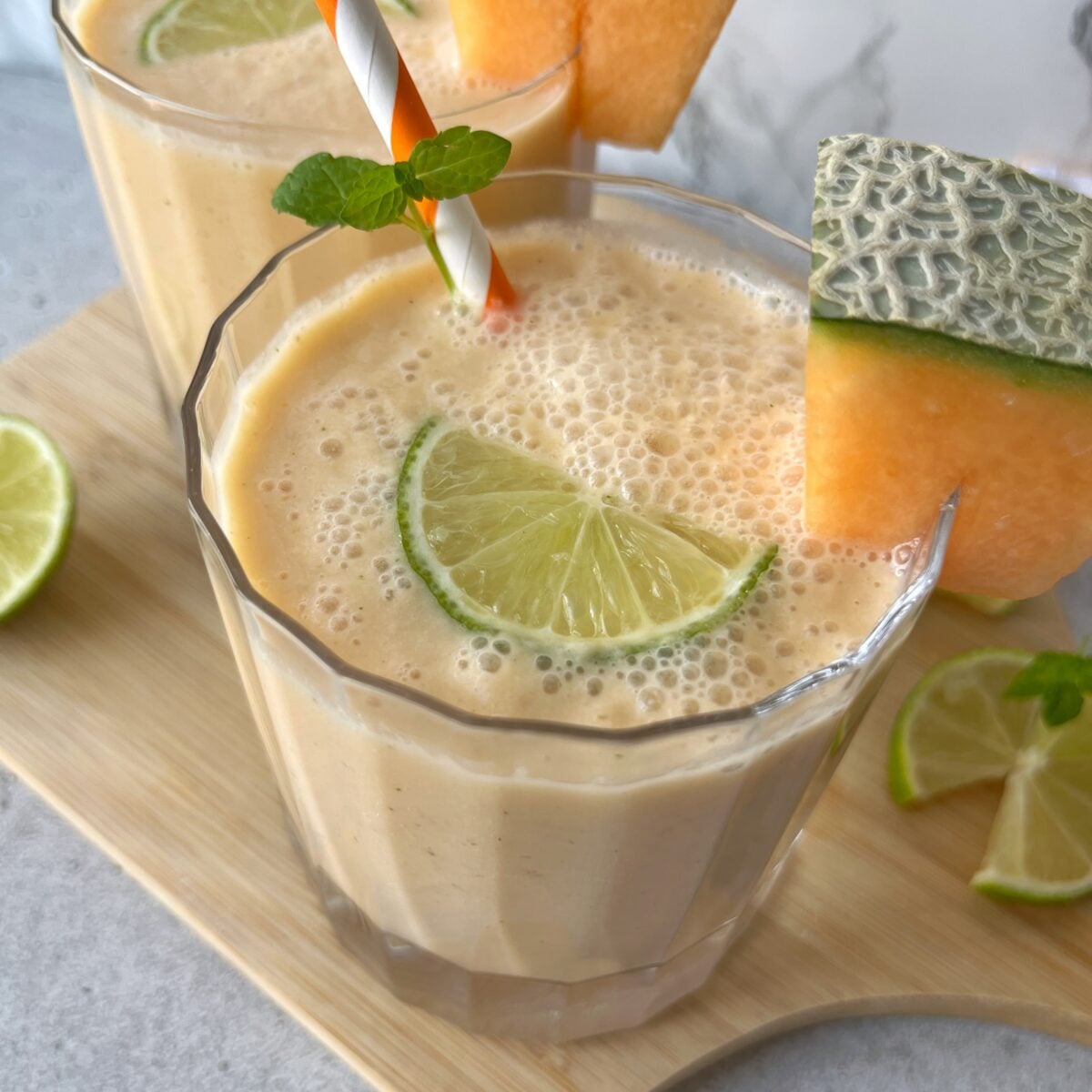 A closeup of a glass of cantaloupe smoothie garnished with a lime slice, a cantaloupe wedge, a mint sprig, and a striped straw.