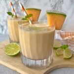 Two glasses of cantaloupe smoothie garnished with lime slices, cantaloupe wedges, mint leaves, and striped straws on a wooden board.