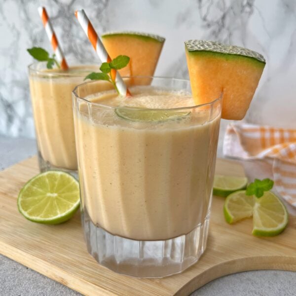 Two glasses of cantaloupe smoothie garnished with lime slices, cantaloupe wedges, mint leaves, and striped straws on a wooden board.