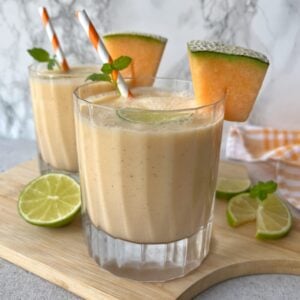 Two glasses of cantaloupe smoothie garnished with lime slices, cantaloupe wedges, mint leaves, and striped straws on a wooden board.