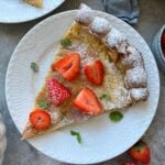 A slice of Dutch baby pancake topped with powdered sugar, fresh strawberry halves, and mint leaves rests on a white plate.