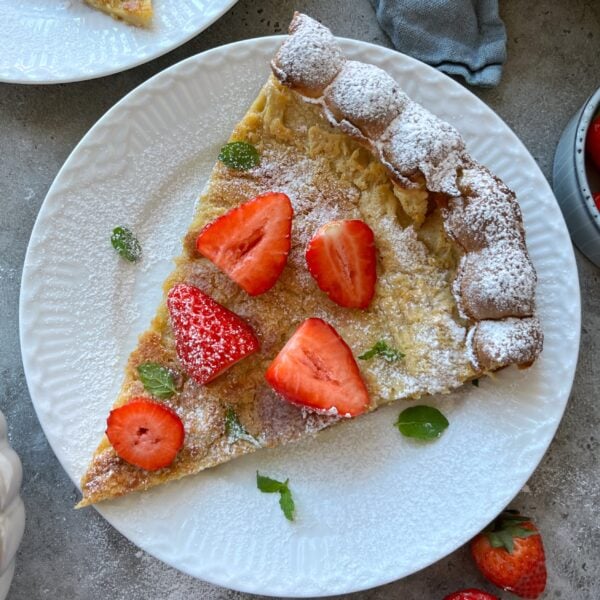 A slice of Dutch baby pancake topped with powdered sugar, fresh strawberry halves, and mint leaves rests on a white plate.