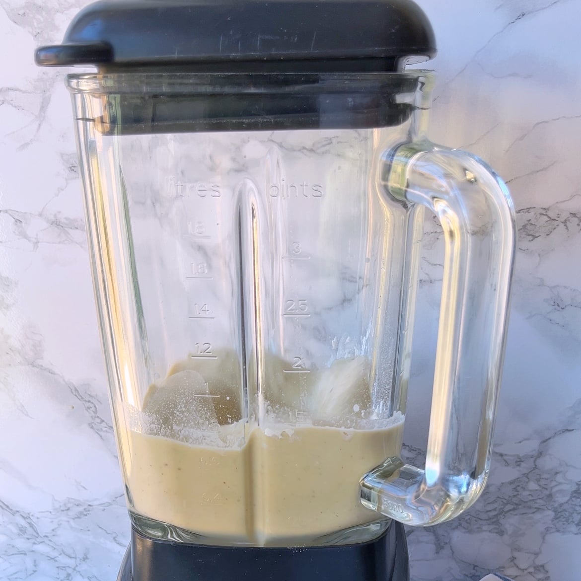A blender containing a partially blended light beige German Pancake mixture sits on a countertop with a marble-patterned background.