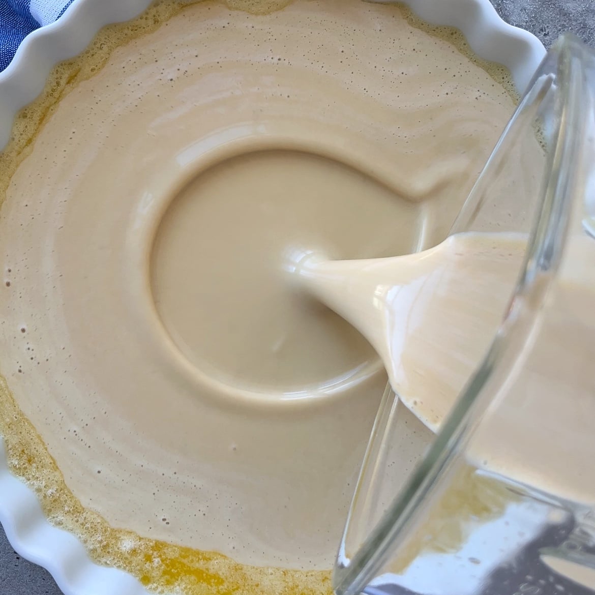 A creamy batter is being poured from a blender into a round, fluted baking dish with melted butter at the bottom.