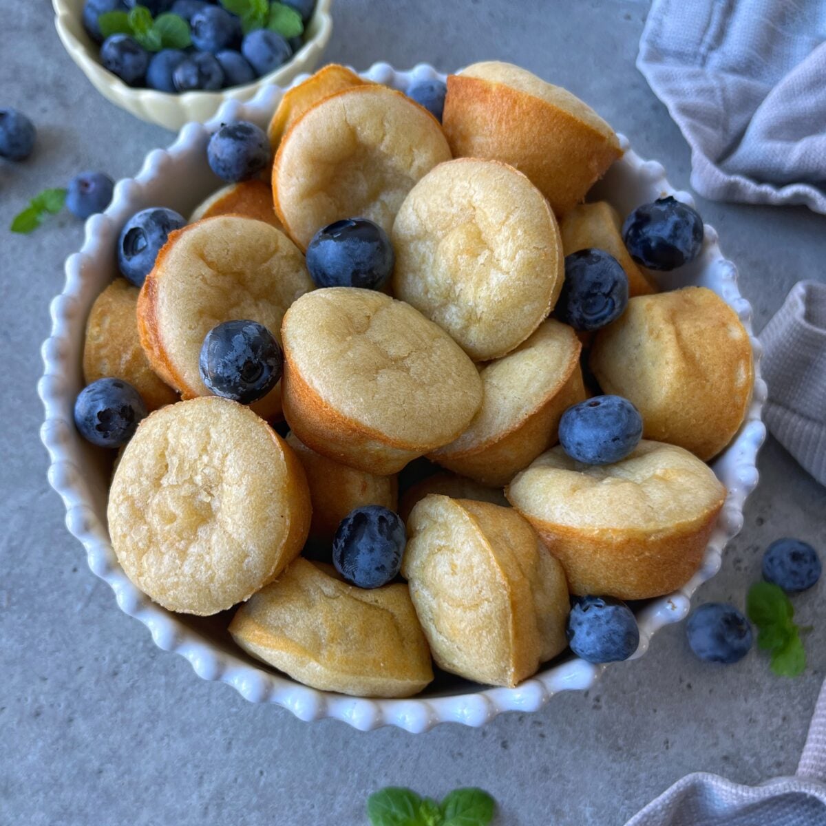 A white bowl filled with small, round pancake muffins and fresh blueberries on a gray surface, with extra blueberries and mint leaves scattered around.