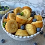 A white bowl filled with golden mini pancakes and fresh blueberries, garnished with mint leaves, sits on a gray countertop.