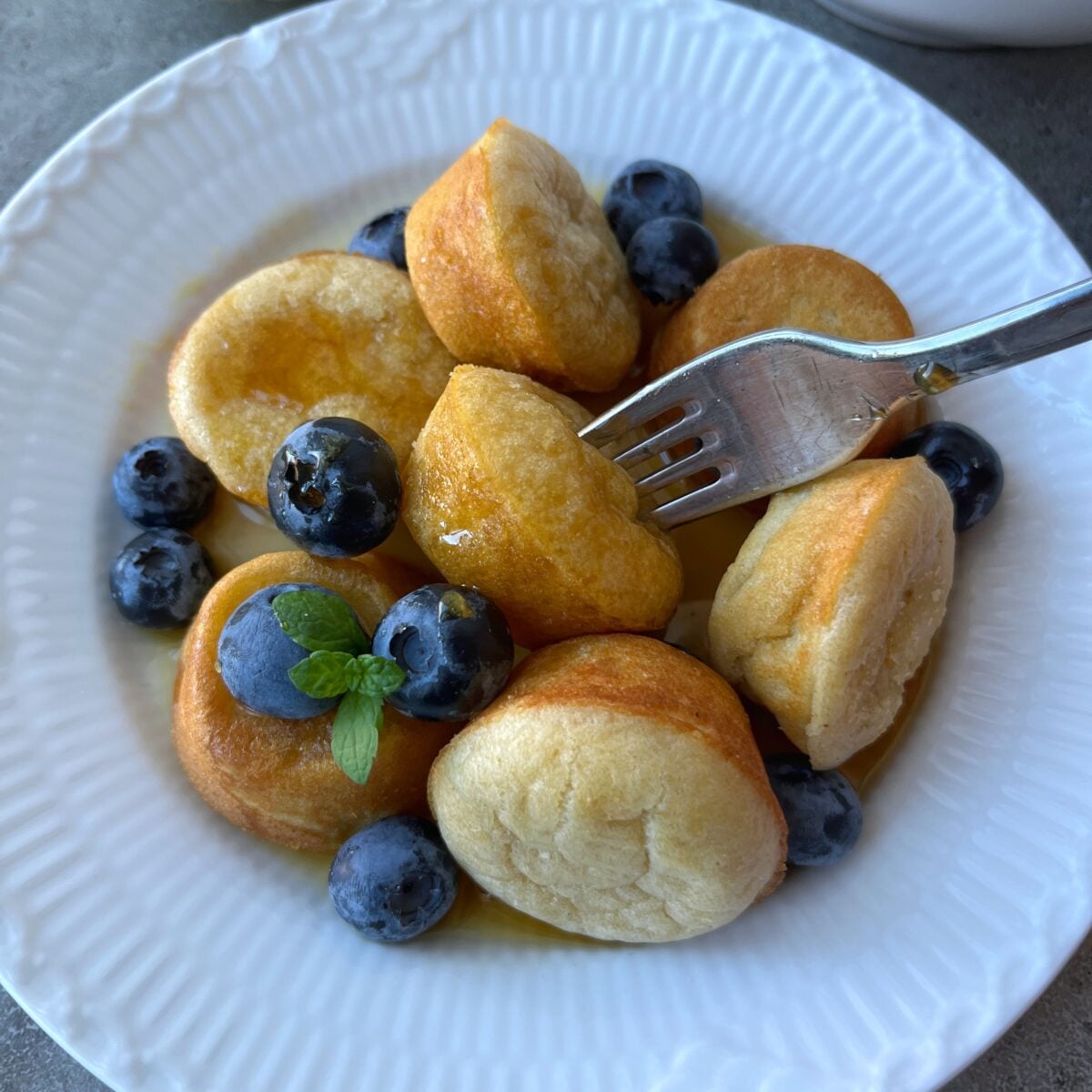 A white plate with small pancake pieces, fresh blueberries, and syrup, featuring a fork holding one Pancake Muffin bite.