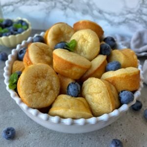 A white bowl filled with golden mini pancakes and fresh blueberries, garnished with mint leaves, sits on a gray countertop.
