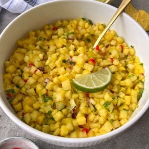 A white bowl filled with Pineapple Mango Salsa, garnished with a lime wedge and mixed with chopped red onion, cilantro, and chili. A gold spoon rests inside the bowl.