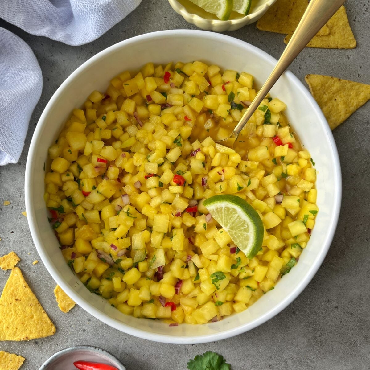 Pineapple Mango Salsa garnished with a lime wedge, with a spoon inside. Tortilla chips, lime slices, and chili peppers are scattered on the gray surface around the bowl.