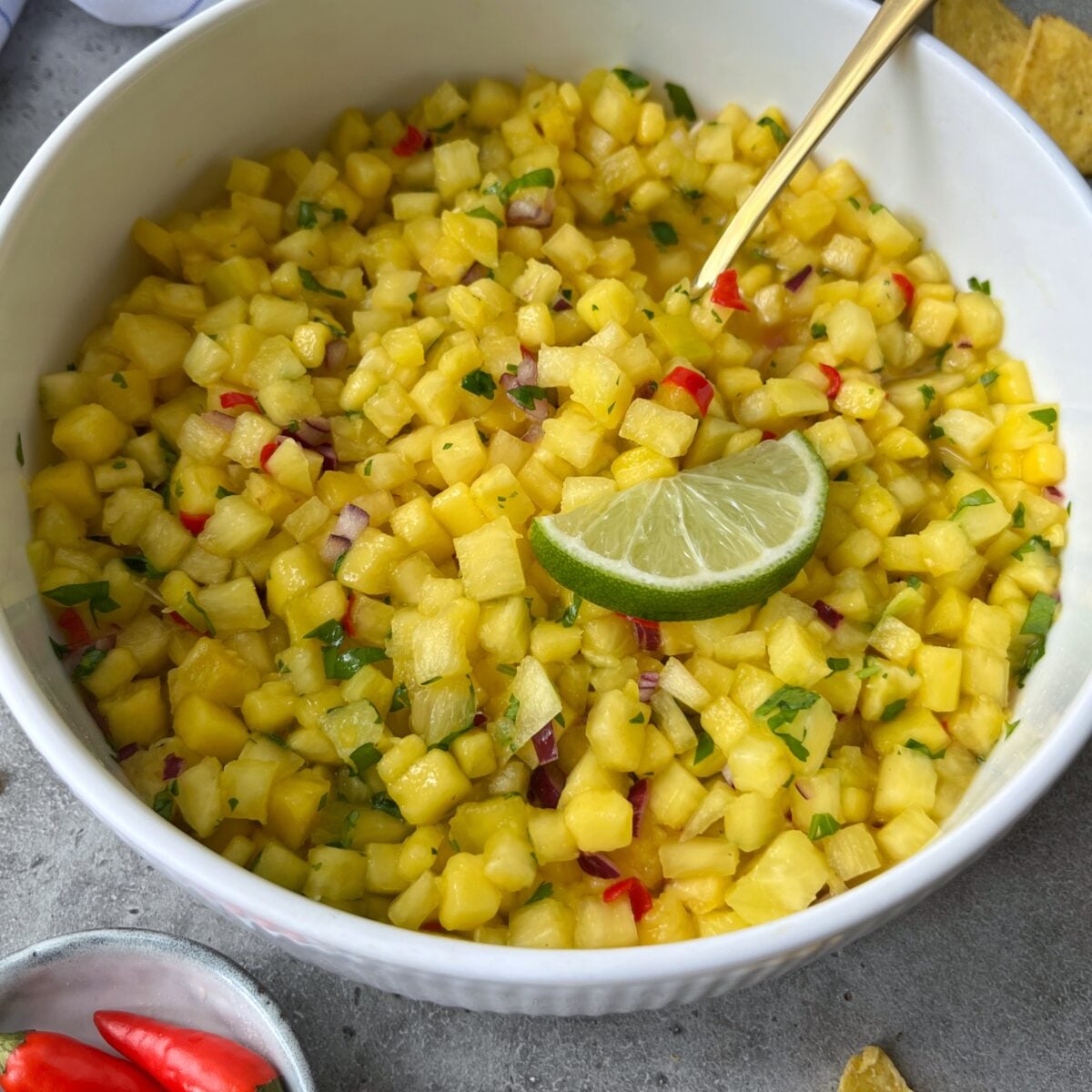 A bowl of Pineapple Mango Salsa with diced pineapple, juicy mango, red onion, red chili, cilantro, and a lime wedge on top. A spoon rests in the vibrant mix.