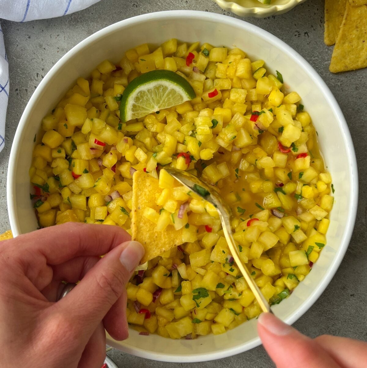 A hand uses a tortilla chip to scoop Pineapple Mango Salsa from a white bowl, garnished with a lime slice.