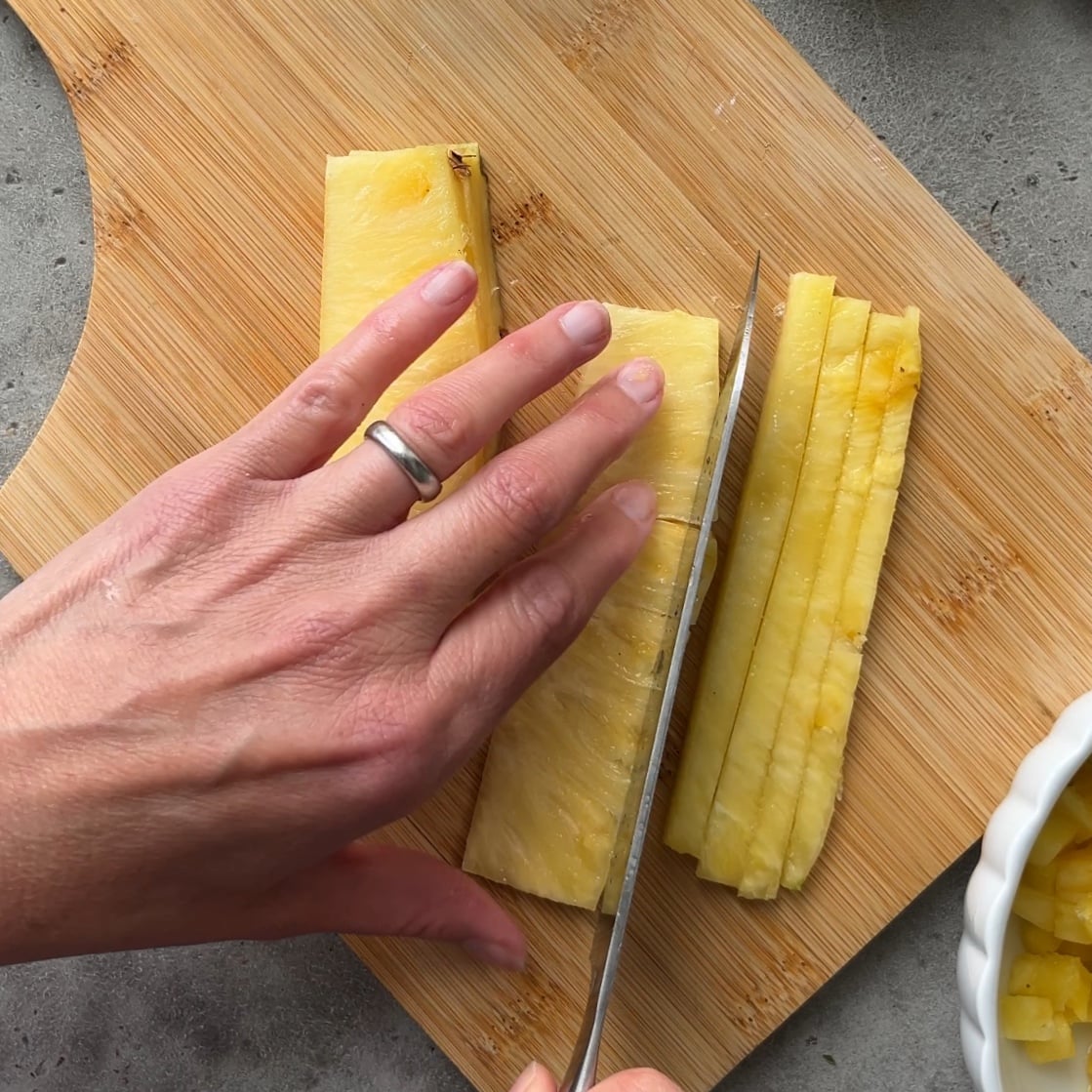 A hand holding a knife slices fresh pineapple on a wooden cutting board.