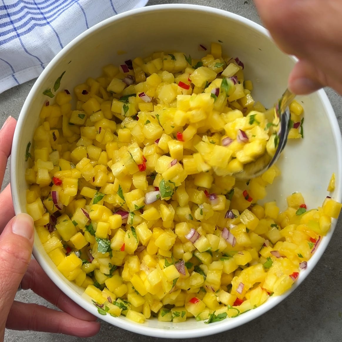 A person mixes a bowl of Pineapple Mango Salsa with diced pineapple and mango, red onion, cilantro, and red chili on a gray surface.