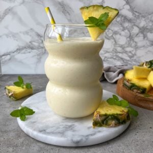 A creamy Pineapple Smoothie in a curvy glass with a striped straw, garnished with a pineapple wedge and mint, sits on a marble slab surrounded by fresh pineapple pieces.