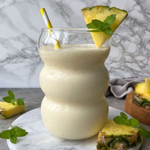 A creamy Pineapple Smoothie in a curvy glass, garnished with a pineapple slice and mint sprig, with a yellow and white striped straw. Pineapple pieces and mint leaves are nearby.