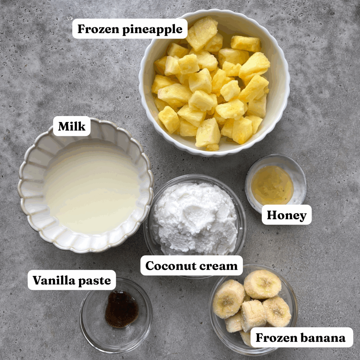 Six labeled ingredients for a Pineapple Smoothie are arranged on a gray surface: frozen pineapple, milk, honey, coconut cream, frozen banana, and vanilla paste.