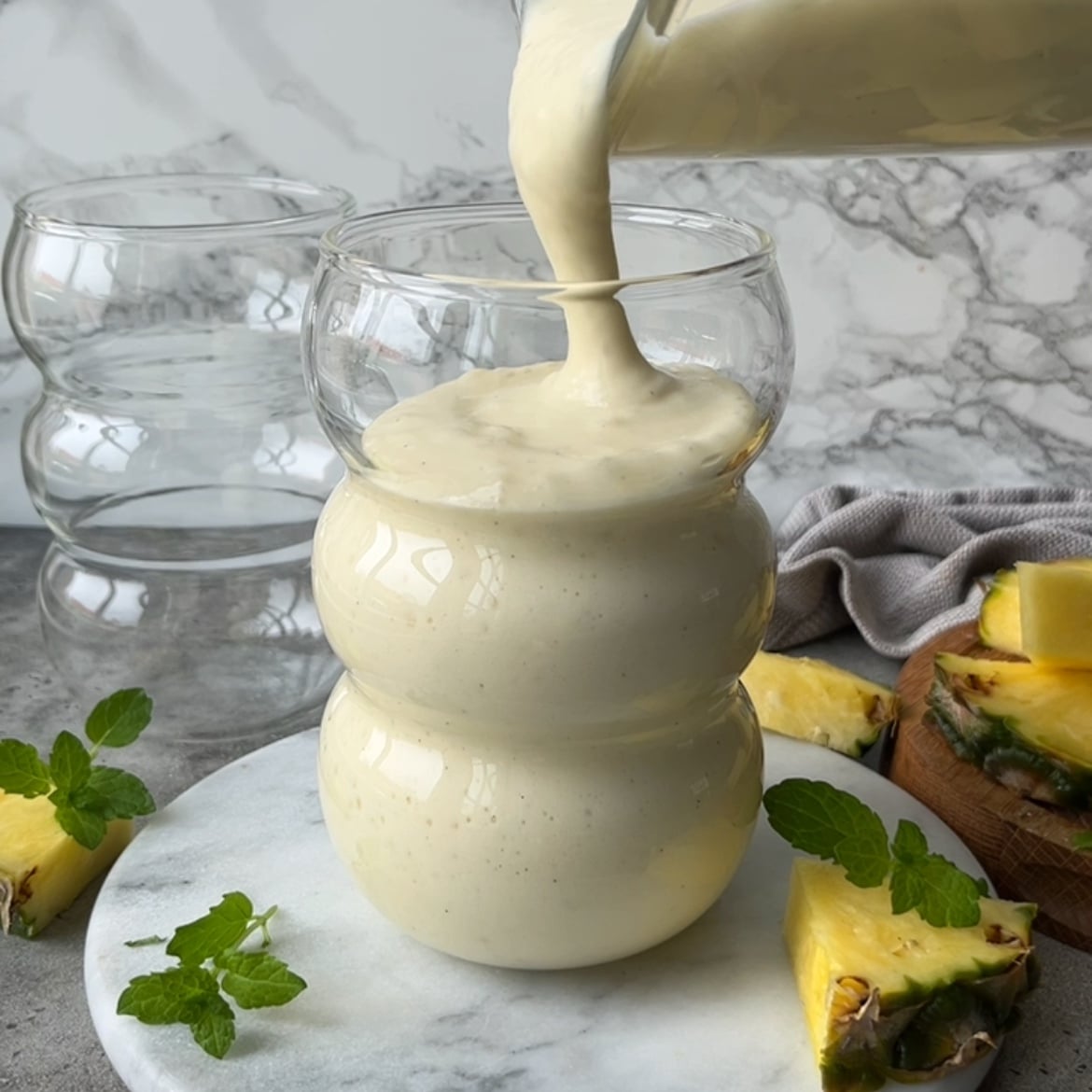 A creamy pineapple smoothie is being poured from a blender into a clear glass, with fresh pineapple slices and mint leaves arranged on a marble surface nearby.