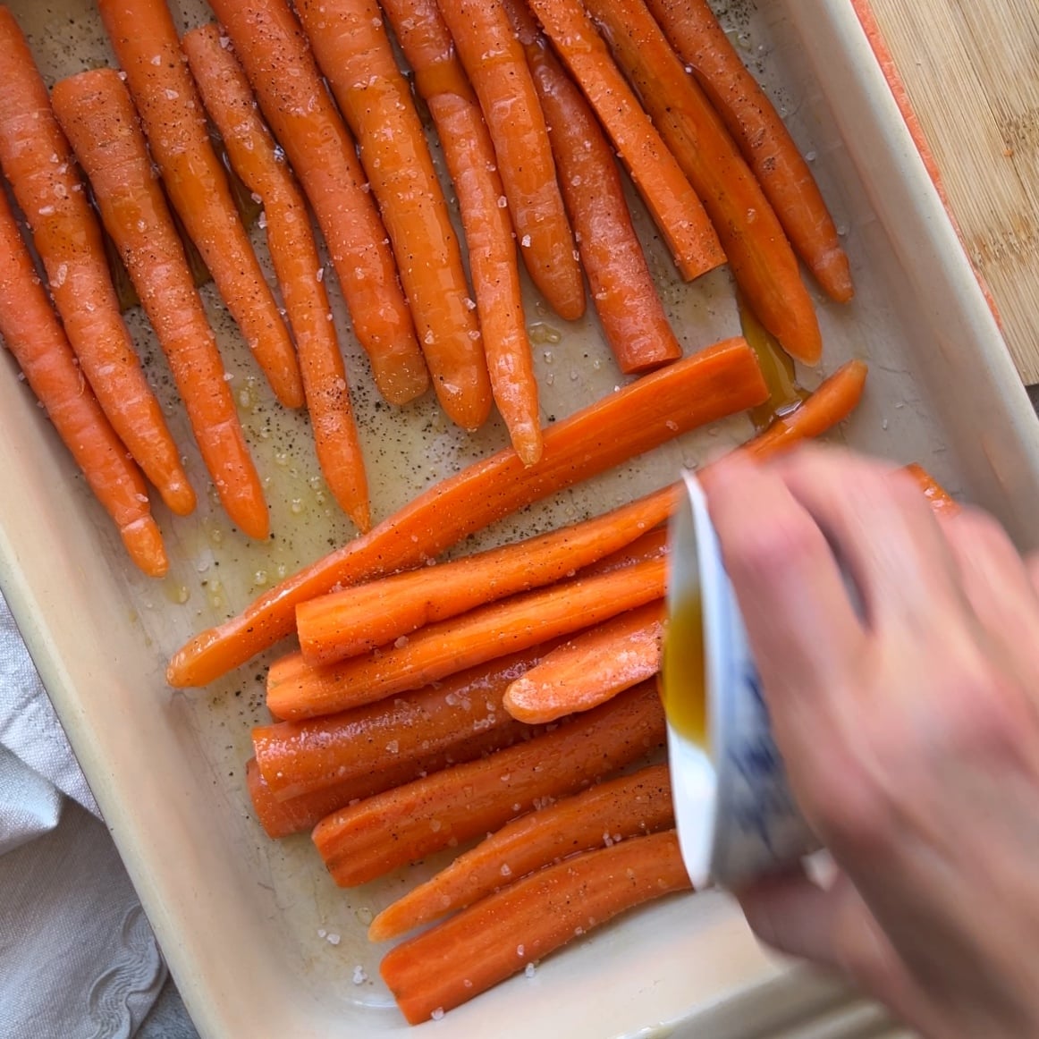 A hand pours maple syrup over raw carrots arranged in a baking dish, sprinkled with salt and pepper.