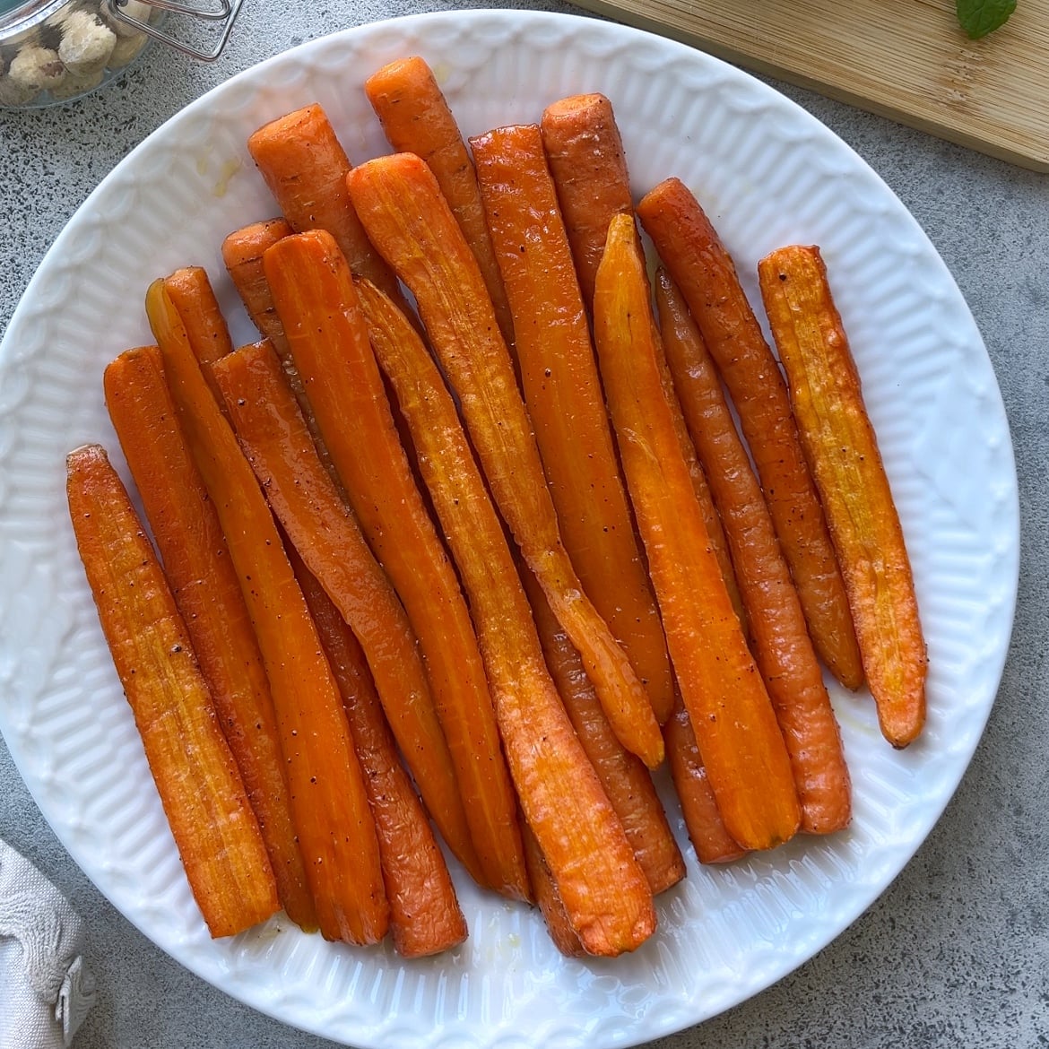 A white plate filled with Roasted Maple Carrots, lightly seasoned and arranged in a single layer on a gray surface.