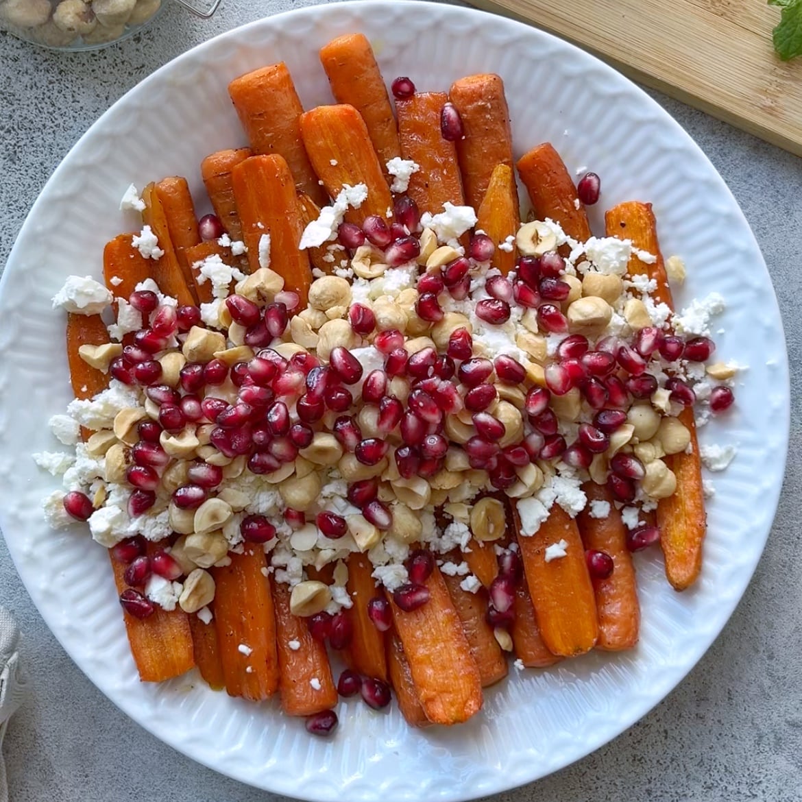 Roasted Maple Carrots on a white plate topped with crumbled feta, pomegranate seeds, and chopped hazelnuts.