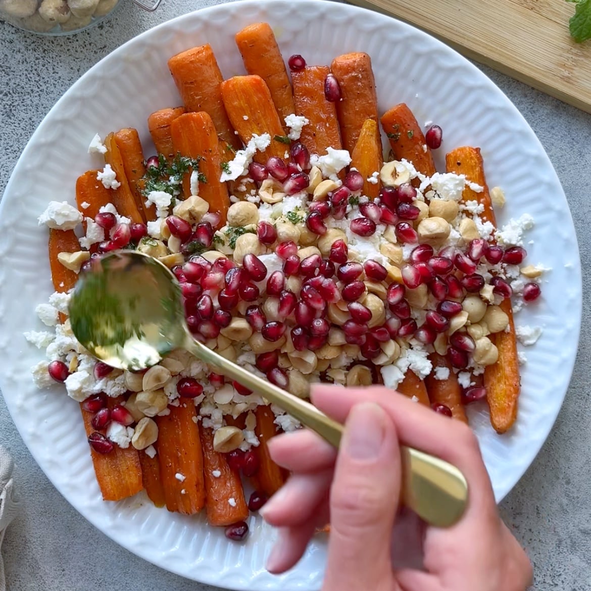 A hand spoons dressing over a plate of Roasted Maple Carrots, topped with feta cheese, hazelnuts, and pomegranate seeds.