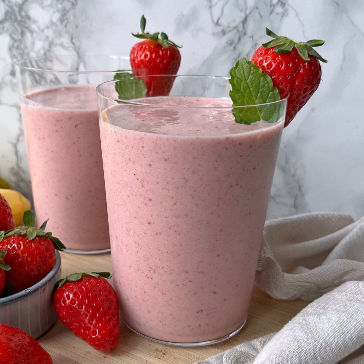 Two glasses of strawberry smoothie garnished with fresh strawberries and mint leaves, set on a wooden surface with extra strawberries and a light-colored cloth nearby.