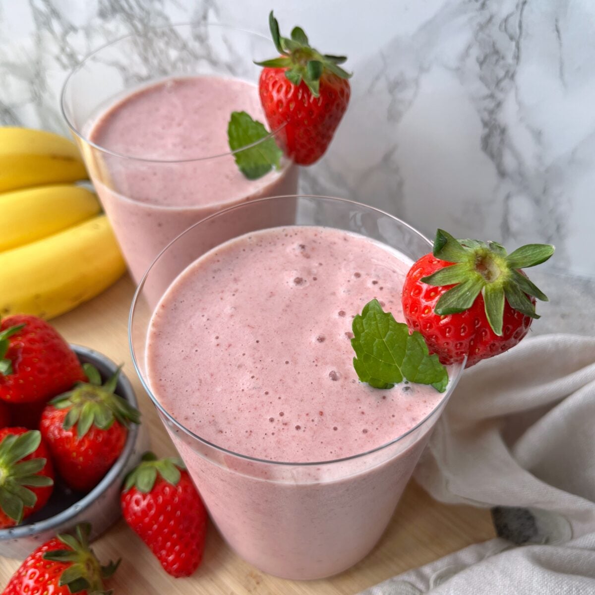 Two glasses of pink strawberry banana smoothie seen from above, each garnished with a fresh strawberry and mint leaf, sit on a wooden surface.