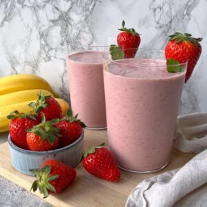 Two glasses of pink strawberry banana smoothie, each garnished with strawberries on the rim, surrounded by fresh strawberries and bananas on a wooden board.