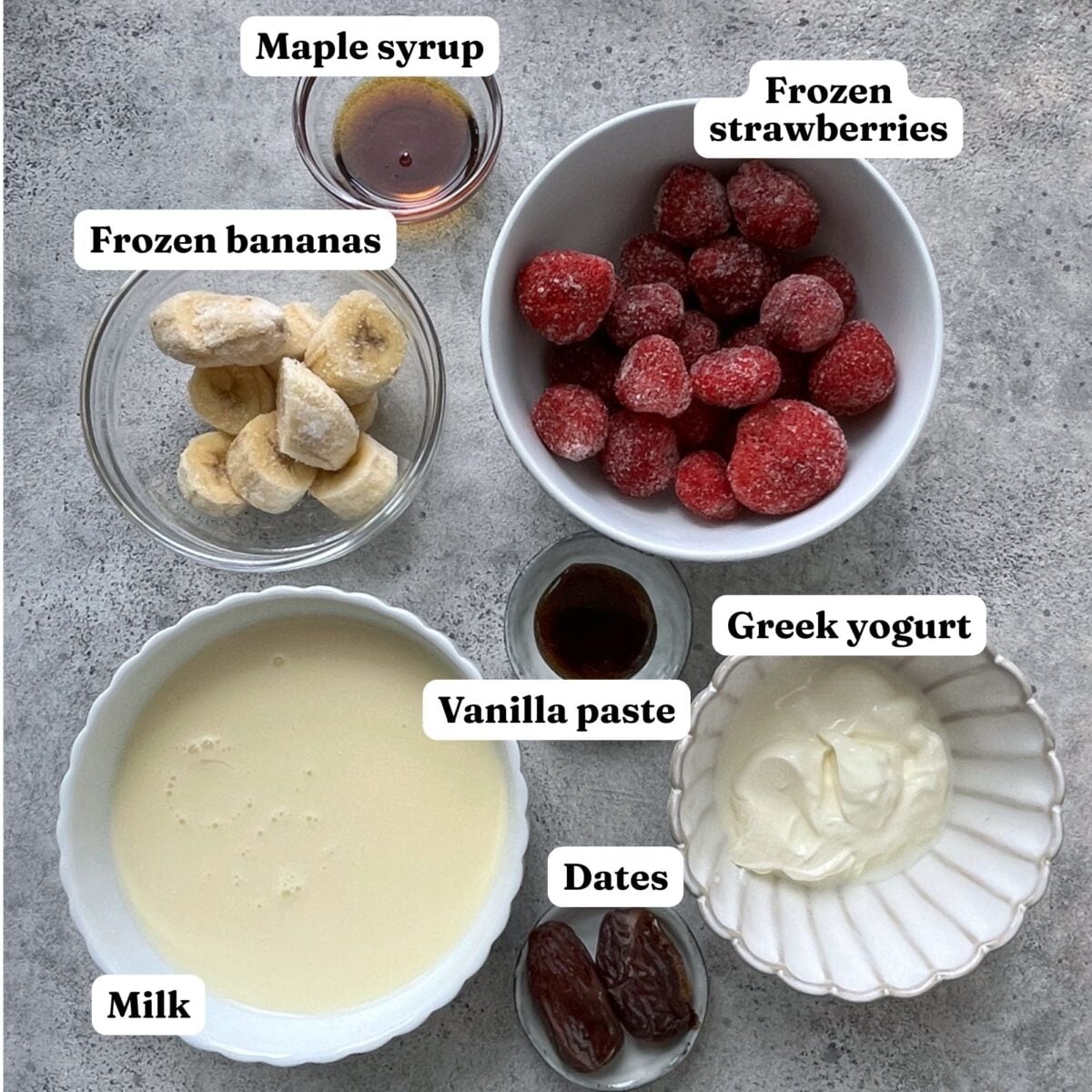 Six labeled bowls and containers display frozen bananas, frozen strawberries, maple syrup, Greek yogurt, dates, vanilla paste, and milk on a gray surface.