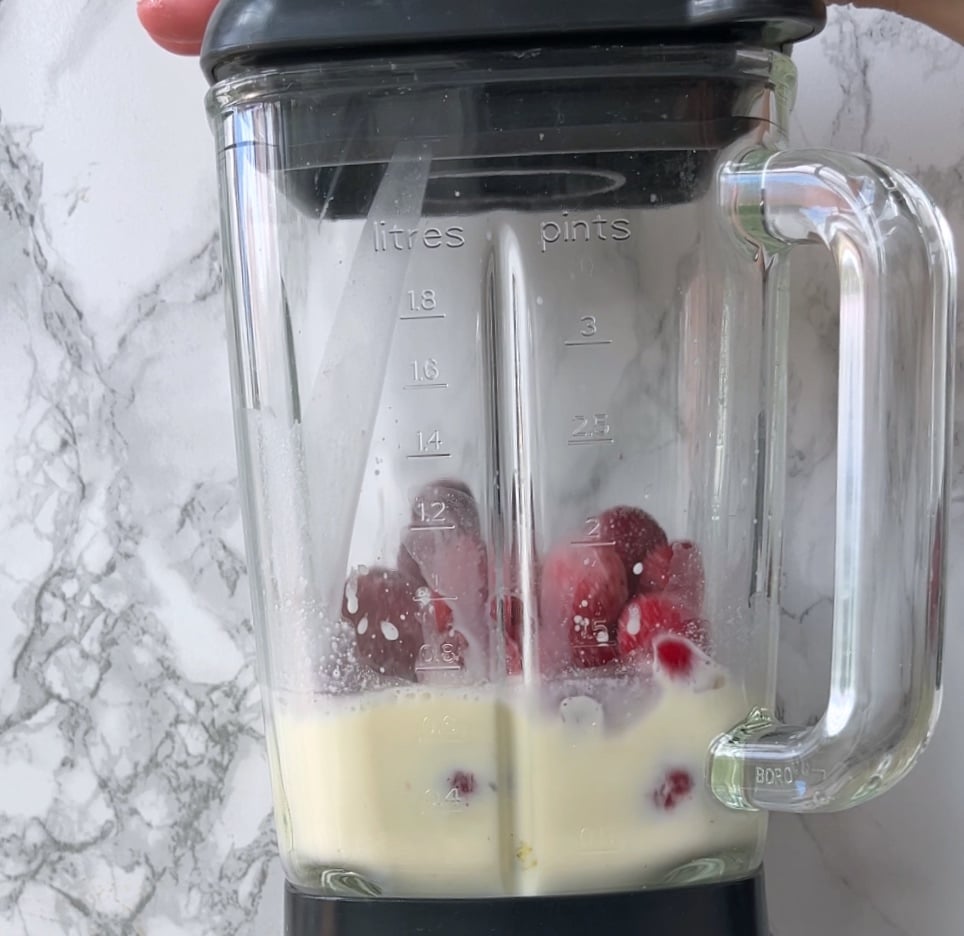 A glass blender contains milk and red strawberries, ready to be blended. The blender has measurement markings in litres and pints.