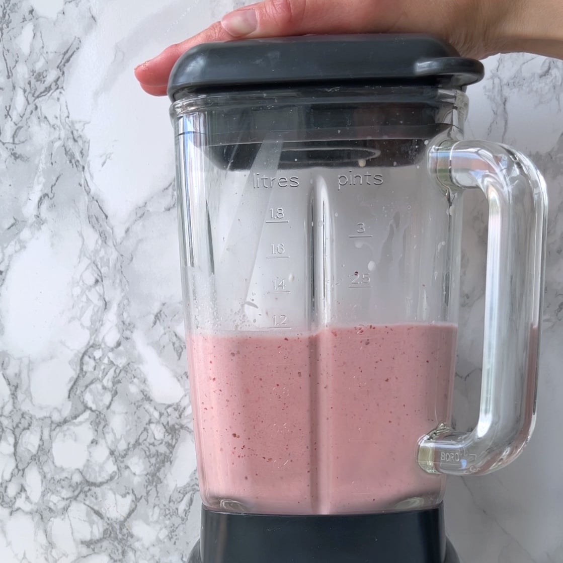 A hand holds the lid of a blender containing a pink smoothie, set on a marble countertop.