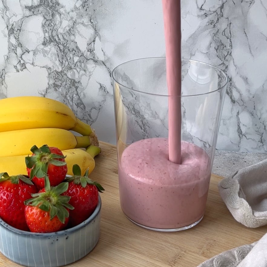 A pink smoothie is being poured into a clear glass, with bananas and strawberries on a wooden surface nearby.