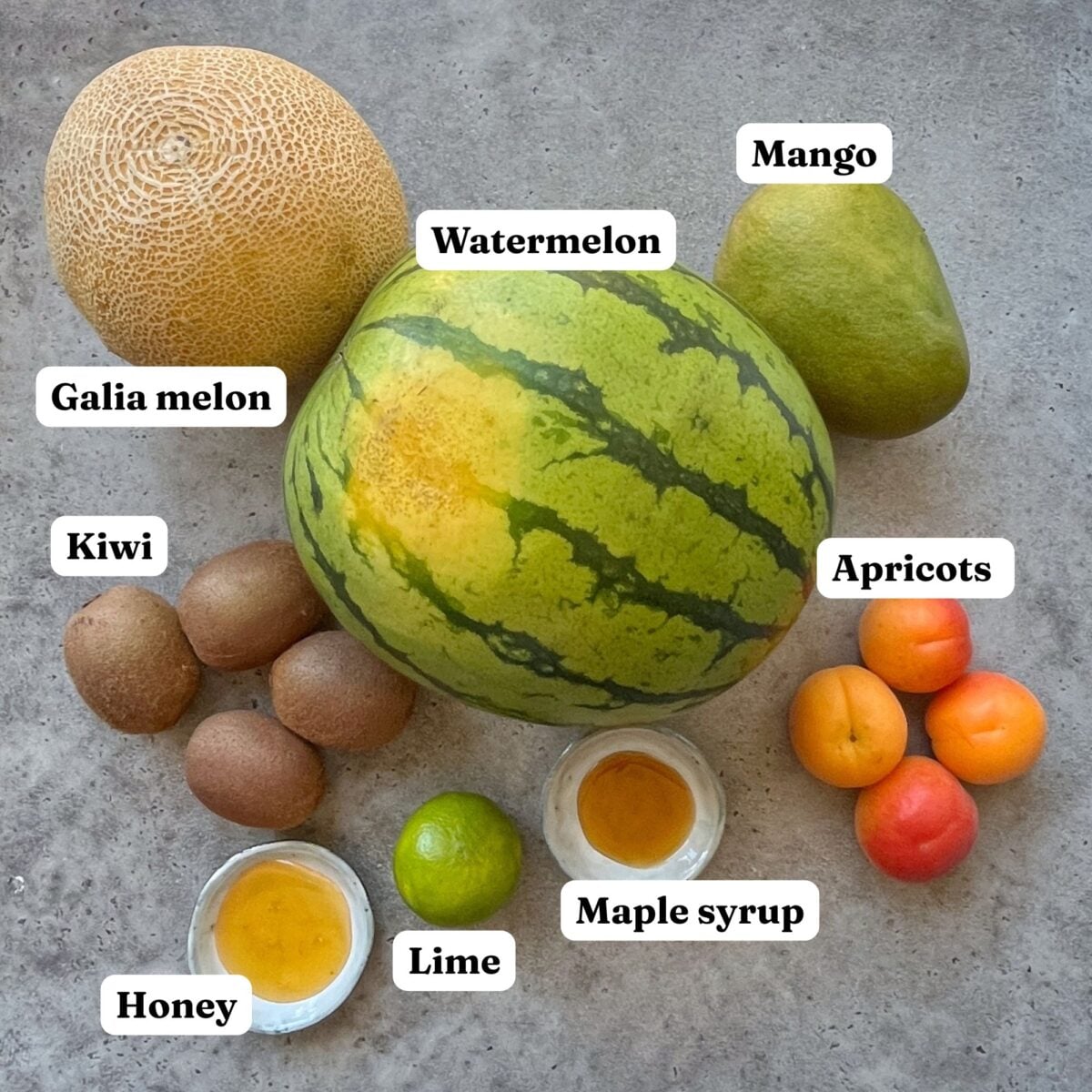 Ingredients needed to make a summer fruit salad including watermelon, Galia melon, apricots, kiwi, mango, lime, honey, and maple syrup.