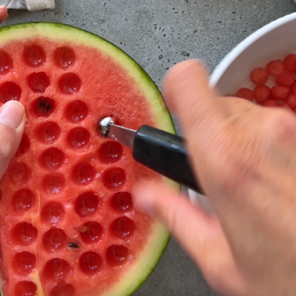 A person uses a melon baller to cut out round balls from a watermelon half.